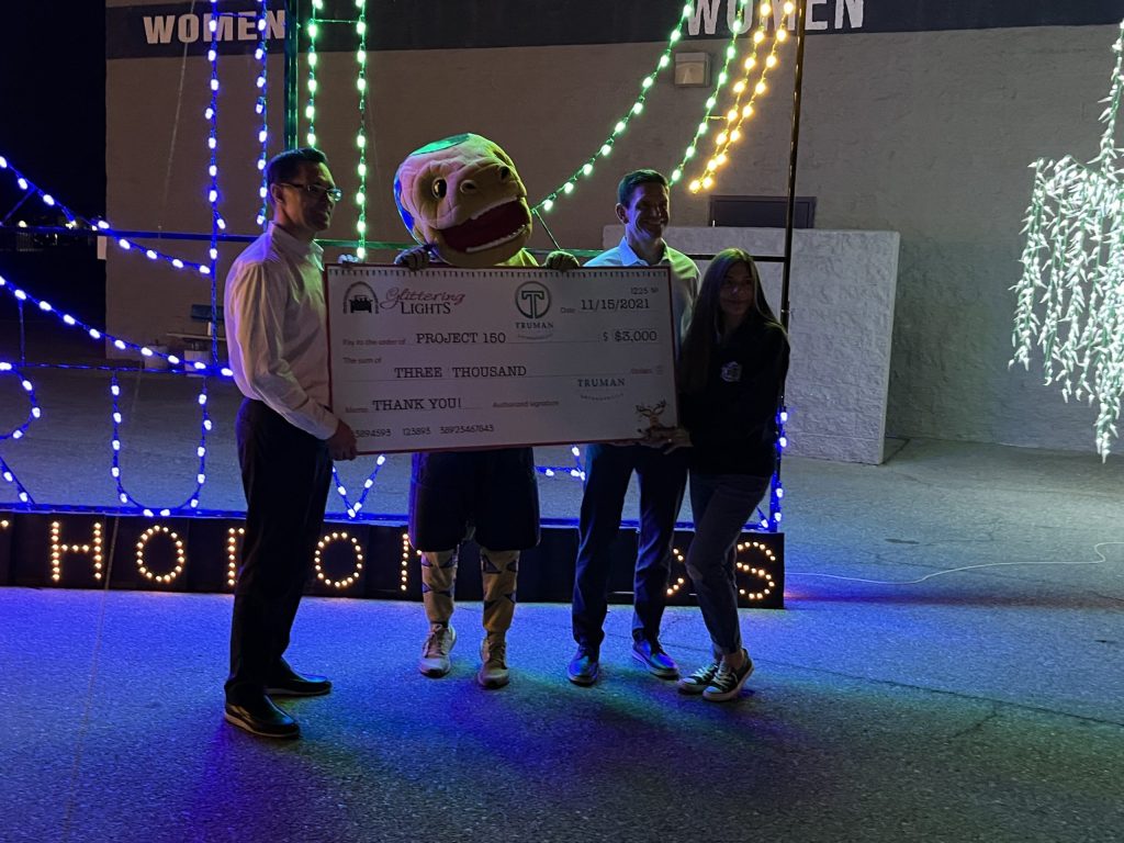 Dr. Mark Truman and Dr. Zach Truman of Truman Orthodontics posing with Chance and Project 150 check at The Neon Forest at Glittering Lights at Las Vegas Motor Speedway